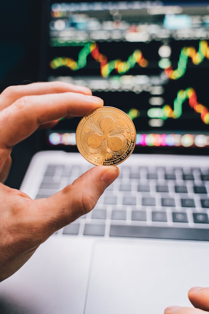 A hand holding a Ripple coin against a laptop screen with trading charts.